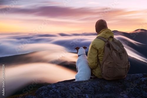 Alone tourist sitting on the edge of the cliff with white dog against the backdrop of an incredible sunrise mountains with flowing fog. Landscape photography