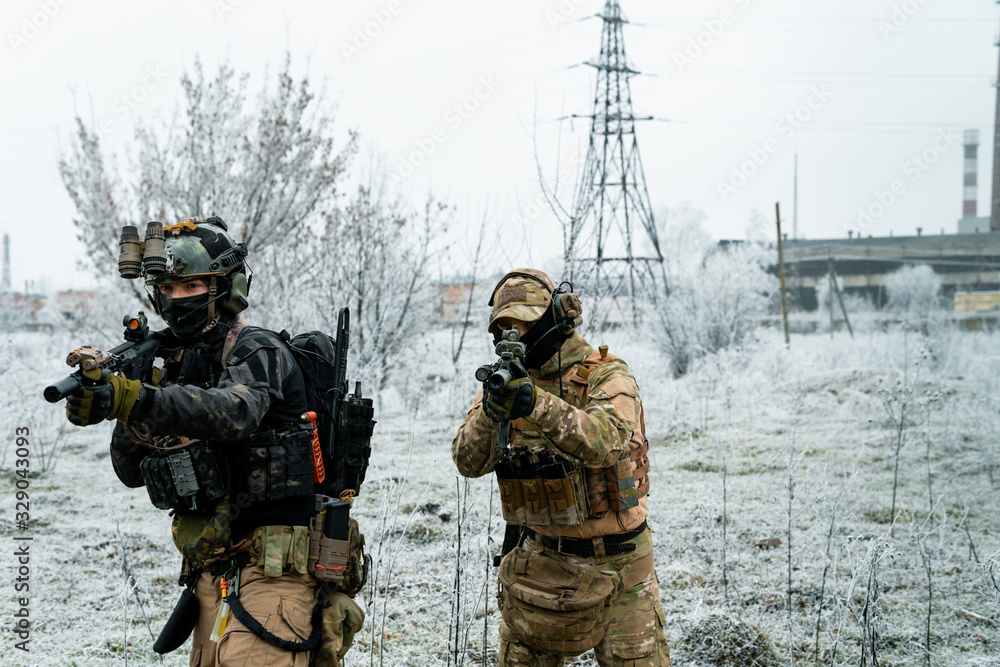 Obraz premium Men in camouflage cloth and black uniform with machineguns with factory on background. Soldiers with muchinegun aims aiming