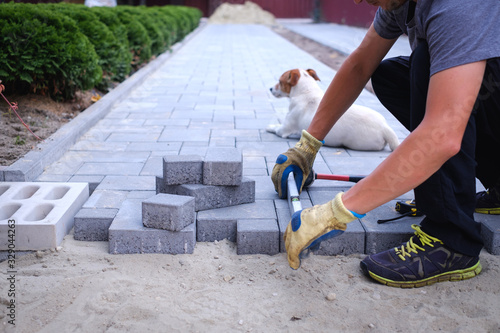 The master in yellow gloves lays paving stones in layers. Garden brick pathway paving by professional paver worker. Laying gray concrete paving slabs in house courtyard on sand foundation base.