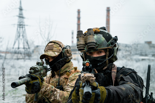 Two men in camouflage cloth and black uniform with machineguns with factory on background. Soldiers with muchinegun aims aiming standing beside wall