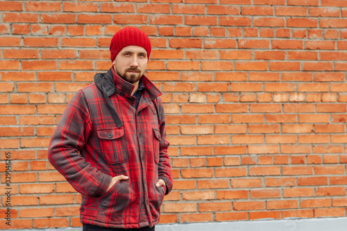 Young bearded man in red coat and hat. Guy with beard is standing in casual clothes against a brick wall and looking at camera.