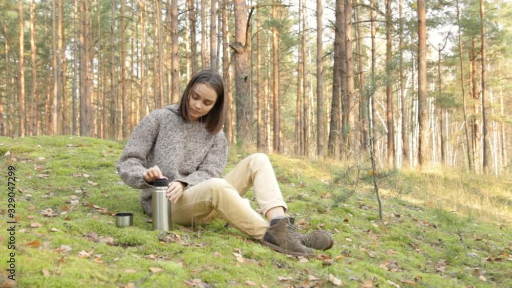 Female hiker drinking from thermos and reading book in forest. Medium shot of pacified female camper sitting on green hill in woods and drinking hot beverage from thermos cup while reading book Female hiker drinking from thermos and reading book in forest. Medium shot of pacified female camper sitting on green hill in woods and drinking hot beverage from thermos cup while reading book