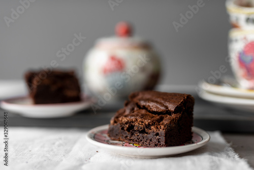 Piece of chocolate brownie on a plate on white linen and wooden table with a tea pot and cups blurry in the back photo