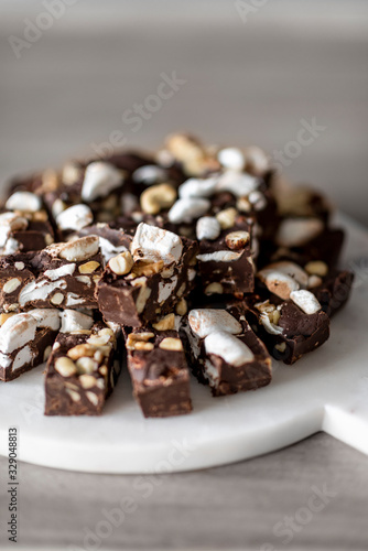 Close up of homemade chocolate chunks topped with nuts and little marshmallows on a wooden and marble round cutting board
