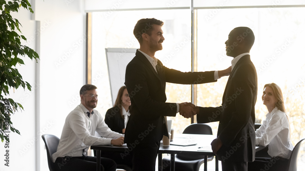 Young male team leader shaking hands with african american employee, congratulating with good job results at diverse business team meeting. Smiling boss rewarding promoting happy mixed race intern. Young male team leader shaking hands with african american employee, congratulating with good job results at diverse business team meeting. Smiling boss rewarding promoting happy mixed race intern.