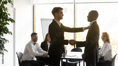 Young male team leader shaking hands with african american employee, congratulating with good job results at diverse business team meeting. Smiling boss rewarding promoting happy mixed race intern.