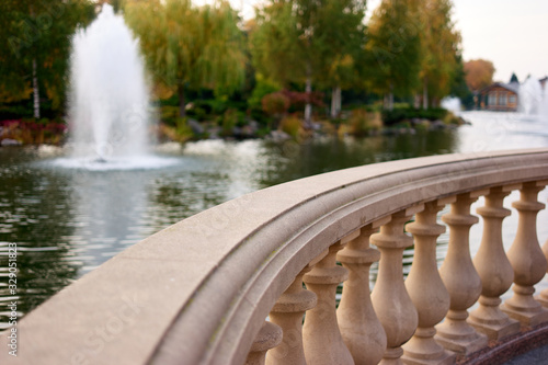 Close up focused stone balustrade railings outdoors. Pond with fountain and trees on background.