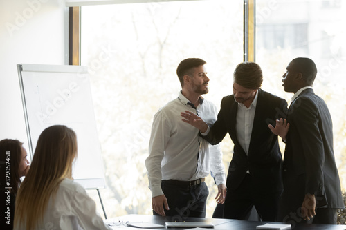 Millennial businessman setting apart two aggressive multiracial coworkers, starting fighting at office. Mixed race young colleagues quarreling shouting at each other, misbehaving during meeting.