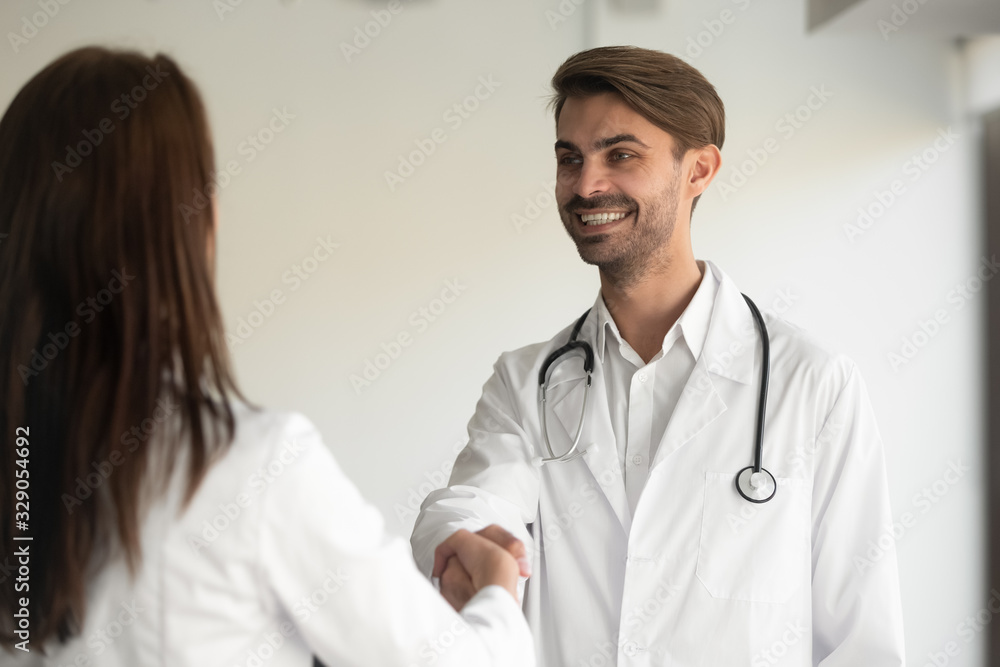 Happy young male general practitioner shaking hands with new female coworker at clinic office. Smiling doctor welcoming nurse at job. Two millennial colleagues thanking each other for cooperation. Happy young male general practitioner shaking hands with new female coworker at clinic office. Smiling doctor welcoming nurse at job. Two millennial colleagues thanking each other for cooperation.