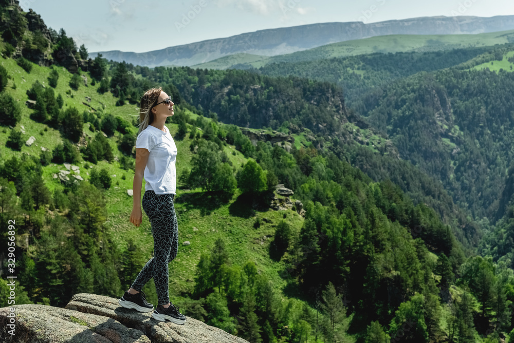 Naklejka premium A beautiful young girl stands high in the mountains and looks at the forest