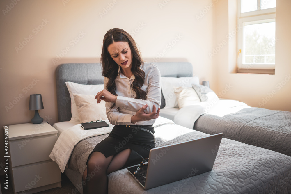 Overworked businesswoman on business trip sitting in hotel room and working on laptop. She is having pain in her arm. Overworked businesswoman on business trip sitting in hotel room and working on laptop. She is having pain in her arm.