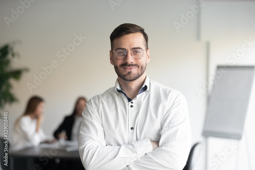 Confident happy young male manager in eyeglasses standing with folded hands head shot portrait. Smiling company owner, startup businessman, team leader, successful employee looking at camera.