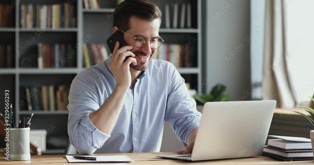 Smiling business man using laptop talking on cell phone sits at desk. Happy confident male professional manager web designer consulting client about online project making business call at workplace.