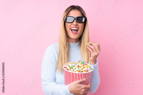 Young Uruguayan woman over isolated pink background with 3d glasses and holding a big bucket of popcorns while pointing front