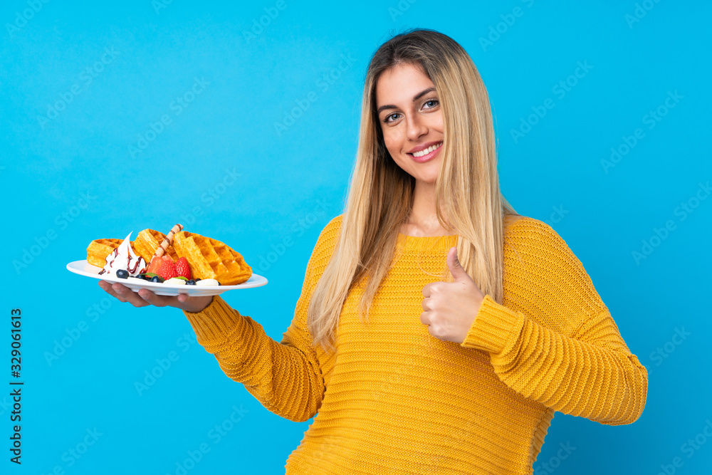 Young woman holding waffles over isolated background with thumbs up because something good has happened Young woman holding waffles over isolated background with thumbs up because something good has happened