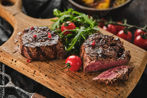 Grilled fillet steaks on wooden cutting board. Succulent thick juicy portions of grilled fillet steak served with tomatoes and roast potatoes on an old wooden board. photo