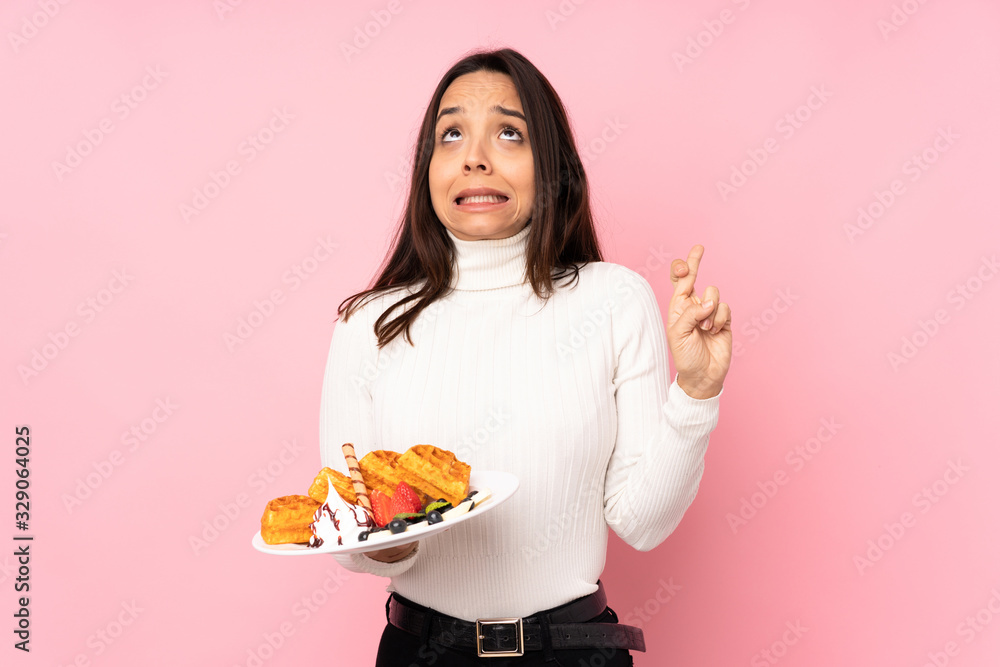 Young brunette woman holding waffles over isolated pink background with fingers crossing and wishing the best Young brunette woman holding waffles over isolated pink background with fingers crossing and wishing the best