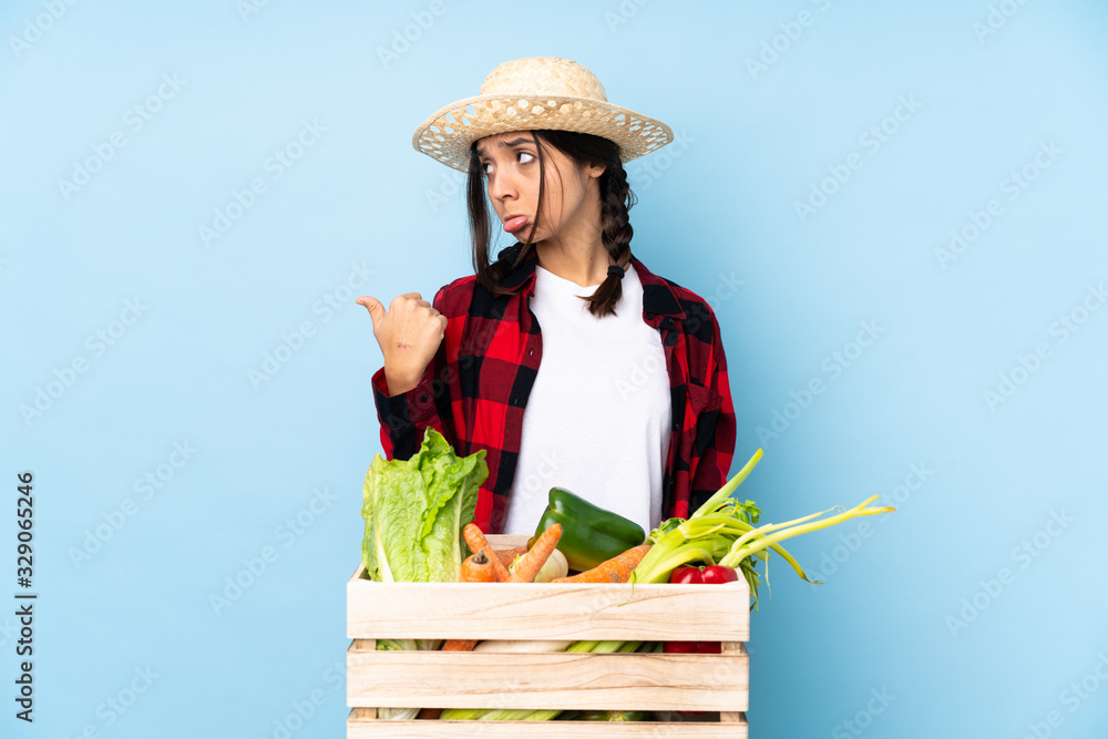 Young farmer Woman holding fresh vegetables in a wooden basket unhappy and pointing to the side