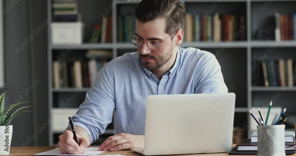Focused business man working on laptop computer making notes sitting at home office desk. Young adult male professional manager doing marketing research writing checklist on paper at workplace.