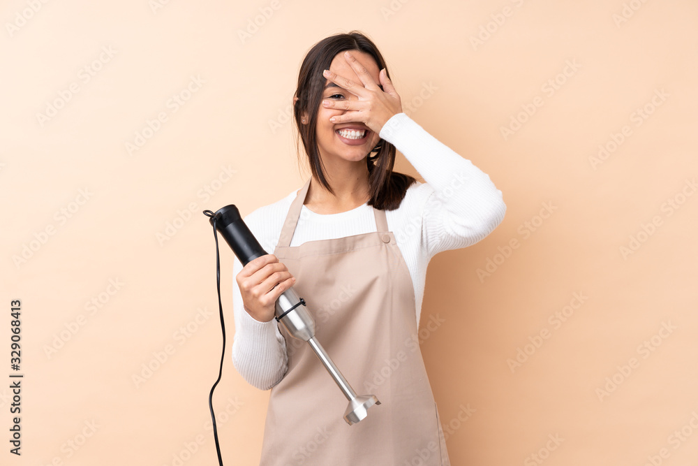 Young brunette girl using hand blender over isolated background covering eyes by hands and smiling Young brunette girl using hand blender over isolated background covering eyes by hands and smiling