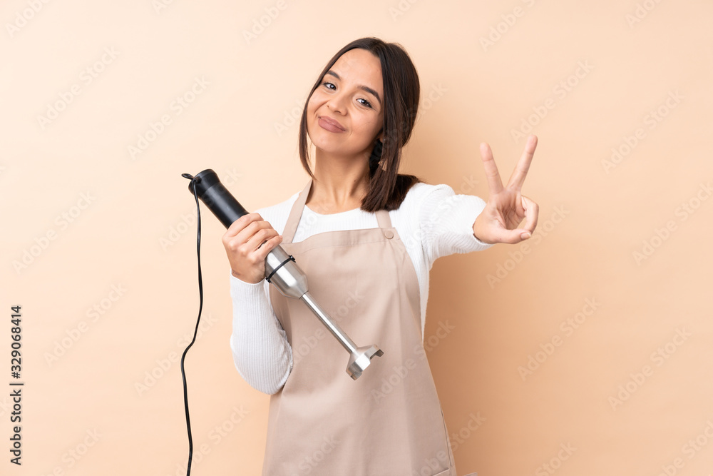 Young brunette girl using hand blender over isolated background smiling and showing victory sign Young brunette girl using hand blender over isolated background smiling and showing victory sign