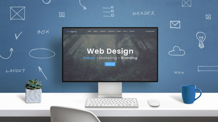 Web design studio home page on modern computer monitor. Office desk with drawings of web design elements on wall