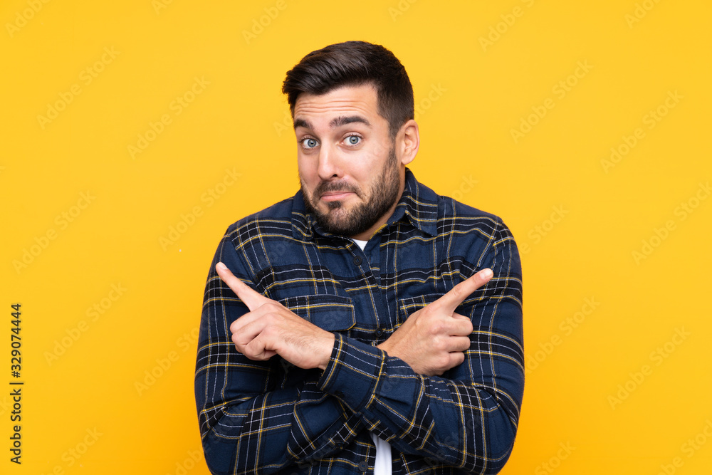 Young handsome man with beard over isolated yellow background pointing to the laterals having doubts Young handsome man with beard over isolated yellow background pointing to the laterals having doubts