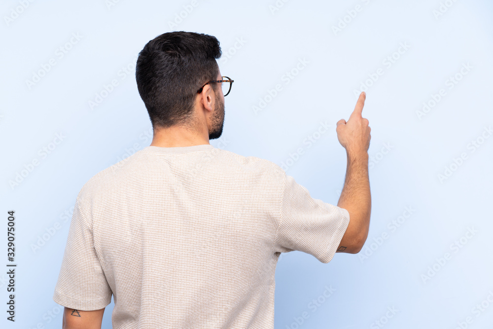 Young handsome man with beard over isolated blue background pointing back with the index finger Young handsome man with beard over isolated blue background pointing back with the index finger