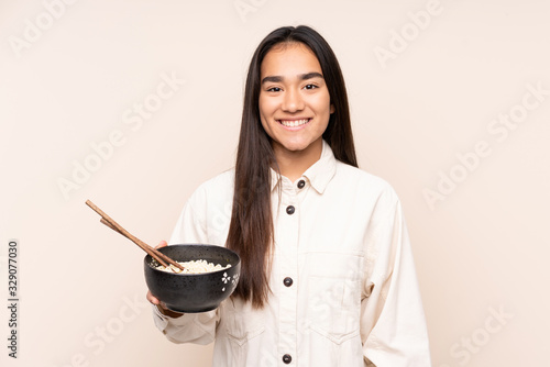 Young Indian woman isolated on beige background smiling a lot while holding a bowl of noodles with chopsticks