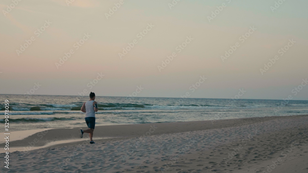 Sporty Hadsome Man is Jogging on Beach with Headphones, Running along Ocean Coast at Sunset or Sunrise Sporty Hadsome Man is Jogging on Beach with Headphones, Running along Ocean Coast at Sunset or Sunrise