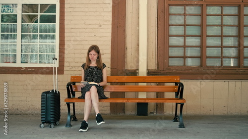 Billede på lærred Caucasian Woman is Waiting for Train on Vacation Sitting on a Bench with Luggage