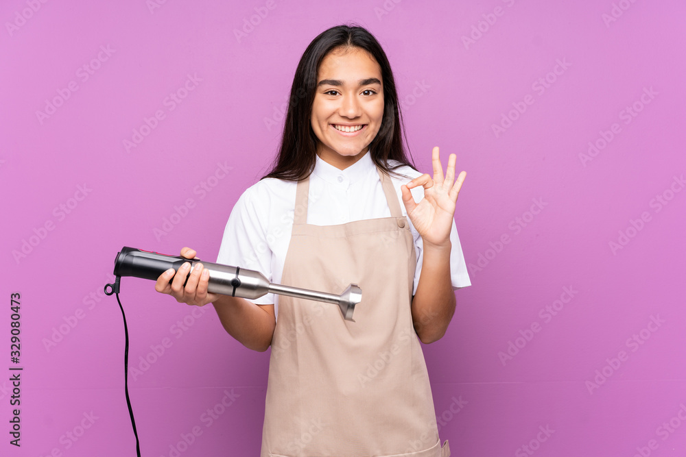 Indian woman using hand blender isolated on purple background showing an ok sign with fingers Indian woman using hand blender isolated on purple background showing an ok sign with fingers