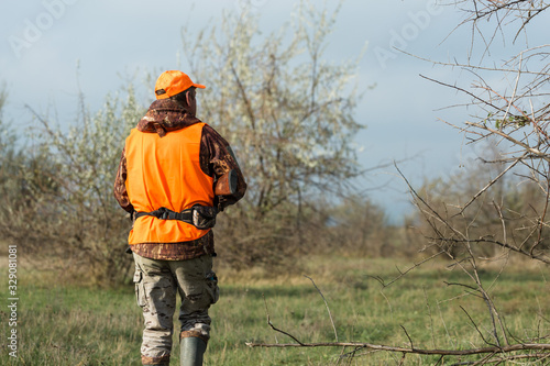 Fotografie, Tablou A man with a gun in his hands and an orange vest on a pheasant hunt in a wooded area in cloudy weather