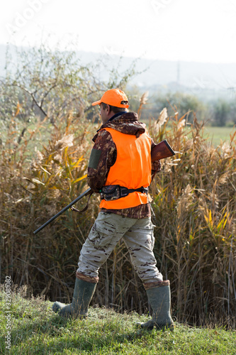 A man with a gun in his hands and an orange vest on a pheasant hunt in a wooded area in cloudy weather. Hunter with dogs in search of game. © Mountains Hunter