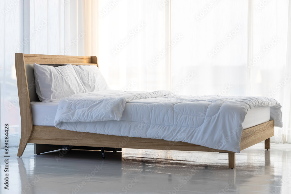 The wooden bed is laid with white sheets in empty room surrounded by white curtains and soft sun light .