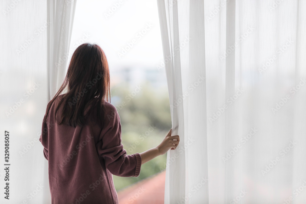 Portrait of beautiful young Asian woman wake up in morning break with freshness and standing near the window and opening white curtains.
