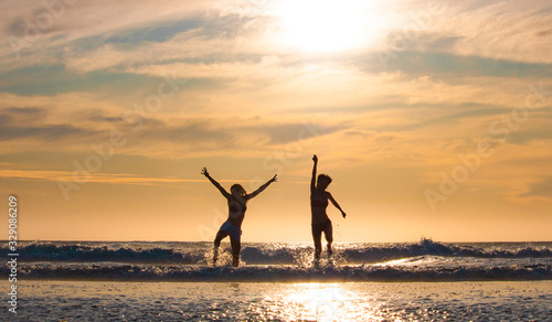 Silhouette of two beautiful women running on a beach shore with waves in the background at sunset or sunrise.