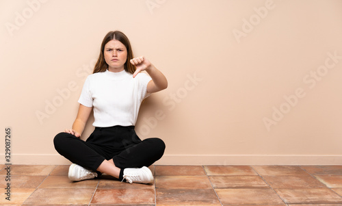Ukrainian teenager girl sitting on the floor showing thumb down with negative expression