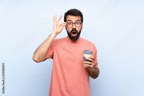 Young man with beard holding a take away coffee over isolated blue background with glasses and surprised