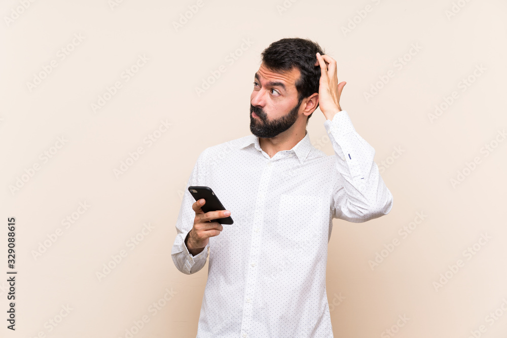 Young man with beard holding a mobile having doubts while scratching head Young man with beard holding a mobile having doubts while scratching head
