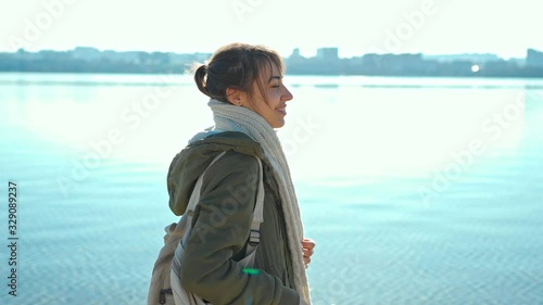 joyful smiling woman in green coat wrappes in white scarf and walks on sandy bay coastline at warm sunny spring day with background of water and cityscape photo