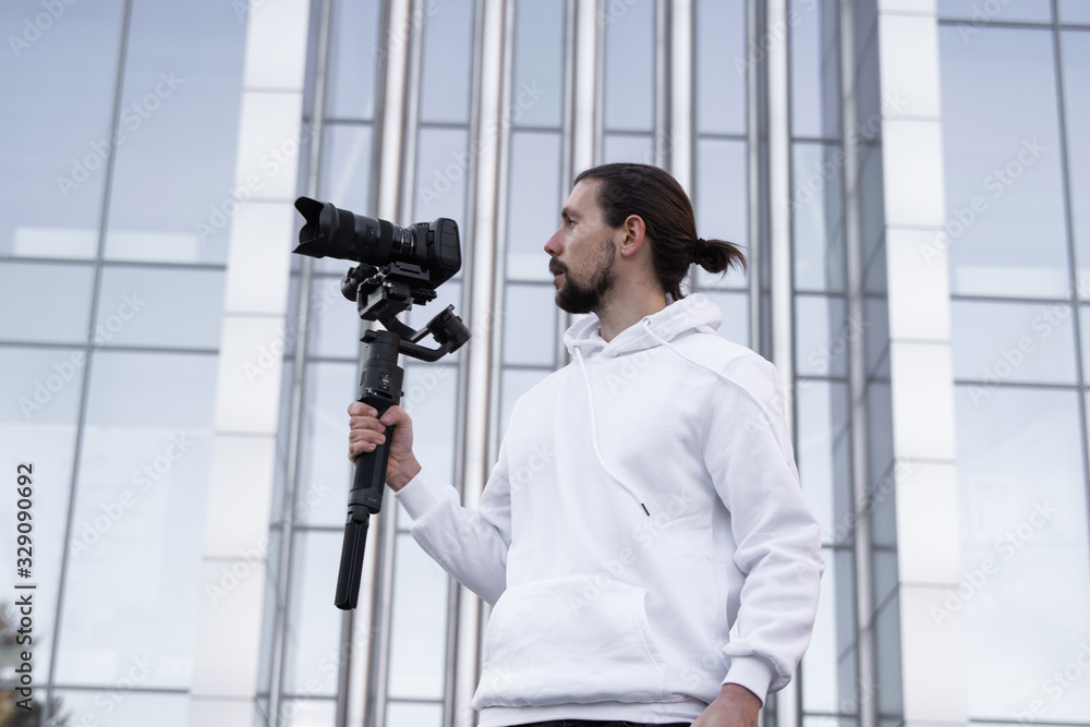 Young Professional videographer holding professional camera on 3-axis gimbal stabilizer. Pro equipment helps to make high quality video without shaking. Cameraman wearing white hoodie making a videos. Young Professional videographer holding professional camera on 3-axis gimbal stabilizer. Pro equipment helps to make high quality video without shaking. Cameraman wearing white hoodie making a videos.