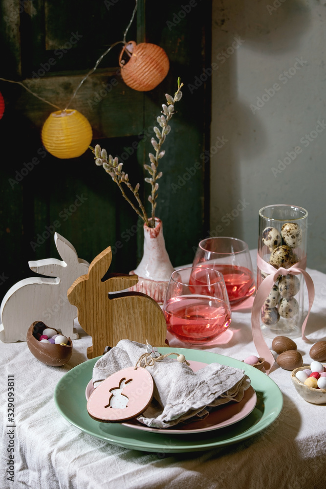 Easter table setting with wooden rabbit decoration, chocolate eggs and sweets, glasses of pink cocktail drinks, empty plates on white tablecloth. Lighting garlands. Holiday dinner.