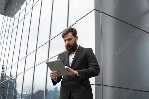 Modern Young bearded Business man working with a digital tablet. Young hipster businessman holding tablet in hands outdoor. Working online with a tablet while standing outside on an office building.