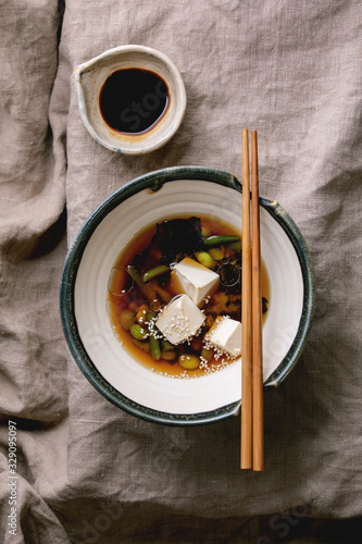 Fototapeta Naklejka Na Ścianę i Meble -  Japanese Miso broth soup with silk tofu cubes, soy beans edamame, green beans in traditional bowl with chopsticks on grey linen table cloth. Asian dinner. Flat lay, space