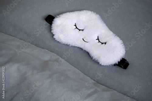 White cute sleep mask made of fluffy faux fur in a cloud shape with closed eyes embroidered on it on a blue grey pillow and blanket. Concept of healthy full sleep and rest