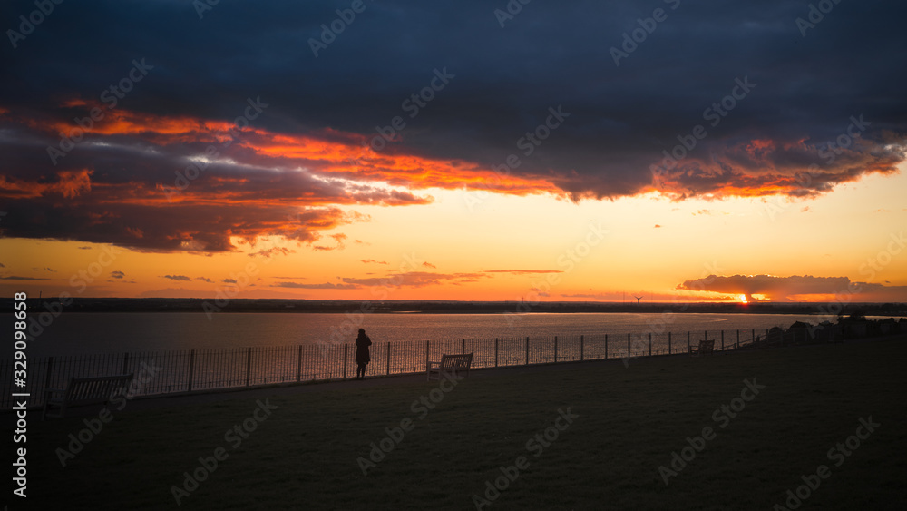 Sunset over the sea. A lady watches from behind a fence with grass in the foreground