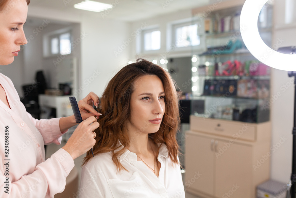 Fototapeta premium The work of the stylist on hairstyles in a bright salon, the girl makes curls and a bunch of beautiful model