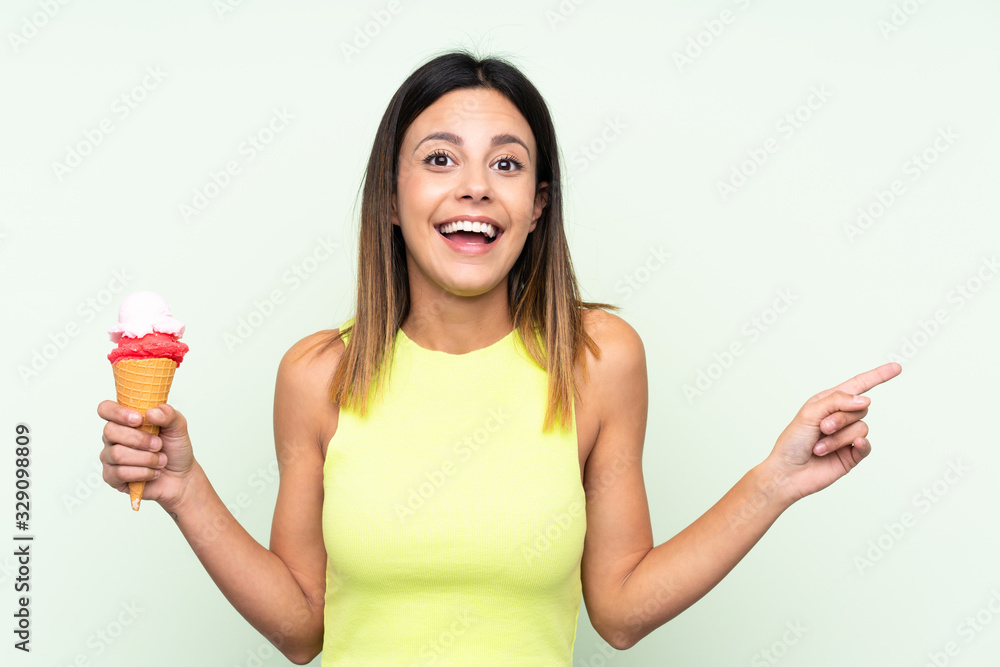 Brunette woman holding a cornet ice cream over isolated green background surprised and pointing finger to the side Brunette woman holding a cornet ice cream over isolated green background surprised and pointing finger to the side