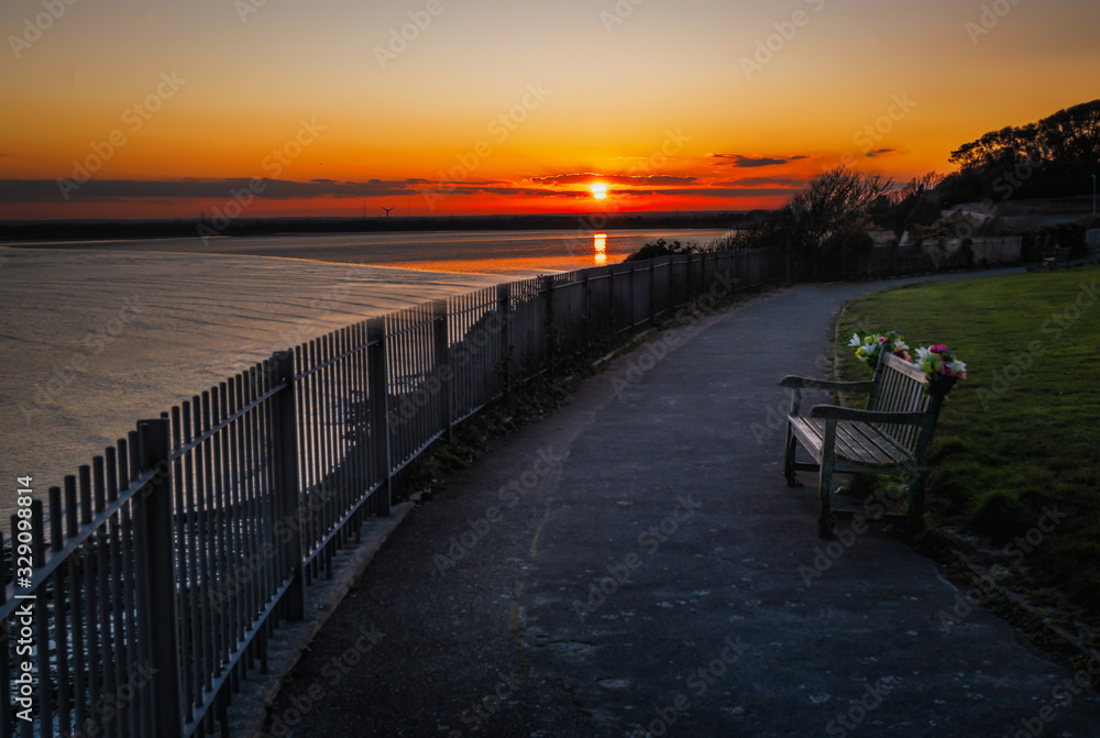 Sunset reflecting in the sea with the cliff top promenade and memorial bench in the foreground in soft focus. Sunset reflecting in the sea with the cliff top promenade and memorial bench in the foreground in soft focus.